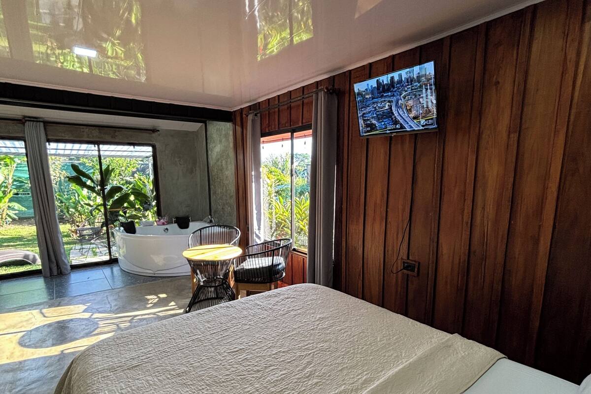 A bedroom with a large bed, a TV mounted on the wall, a small table with chairs, and a view of greenery outside.