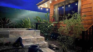 Et træhus med en stentrappe op til en terrasse, omgivet af forskellige planter og et grillområde.