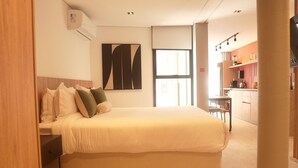 A bedroom with a large bed, a nightstand, a wall-mounted air conditioner, a window, and a kitchenette in the background. - Studio 2 pessoas 36m² - Wi-Fi Rapido e Estacao de Trabalho (Florianópolis)