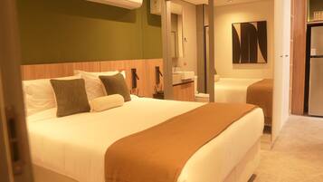 Habitación de hotel con una cama grande, una mesita de noche, un espejo, un lavamanos y una puerta que da a otra habitación.