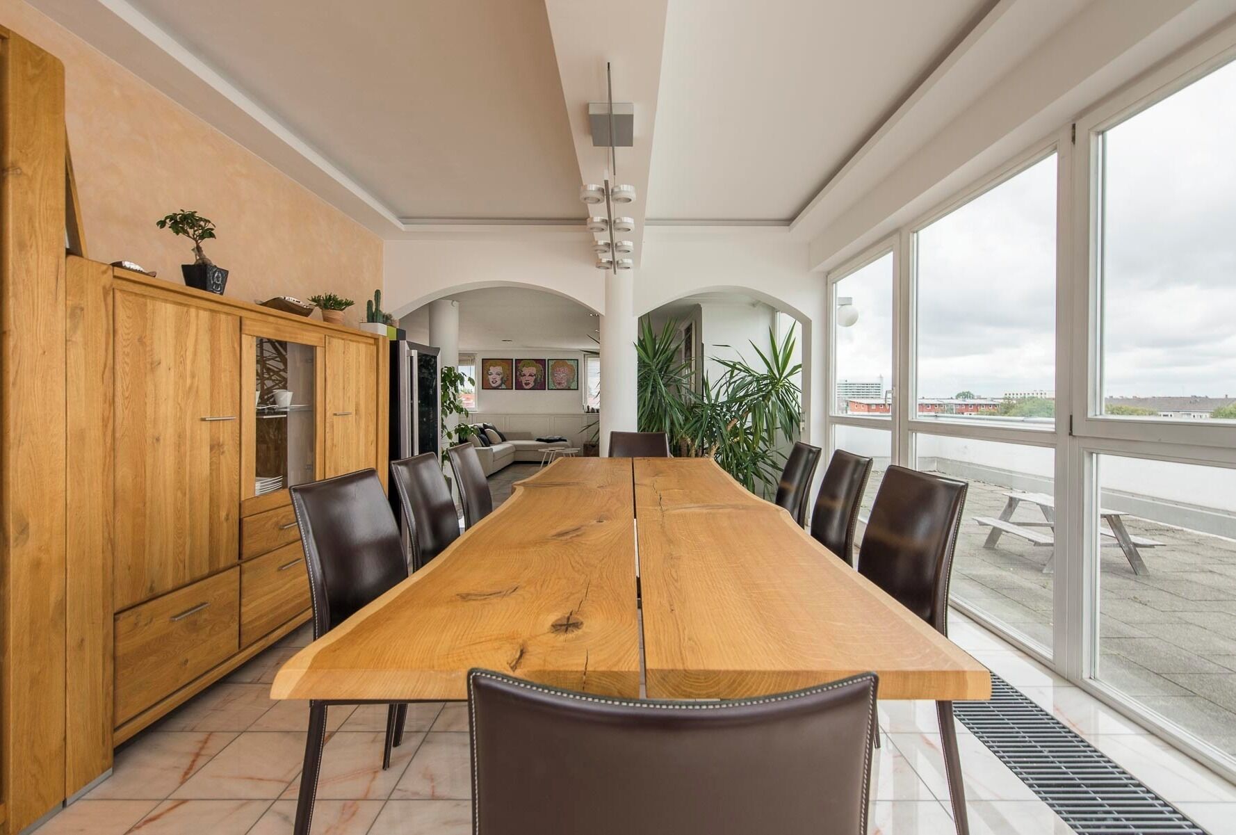 Un comedor amplio con una larga mesa de madera y sillas modernas, ventanas grandes que ofrecen vistas al exterior, y una cocina con armarios de madera y plantas.