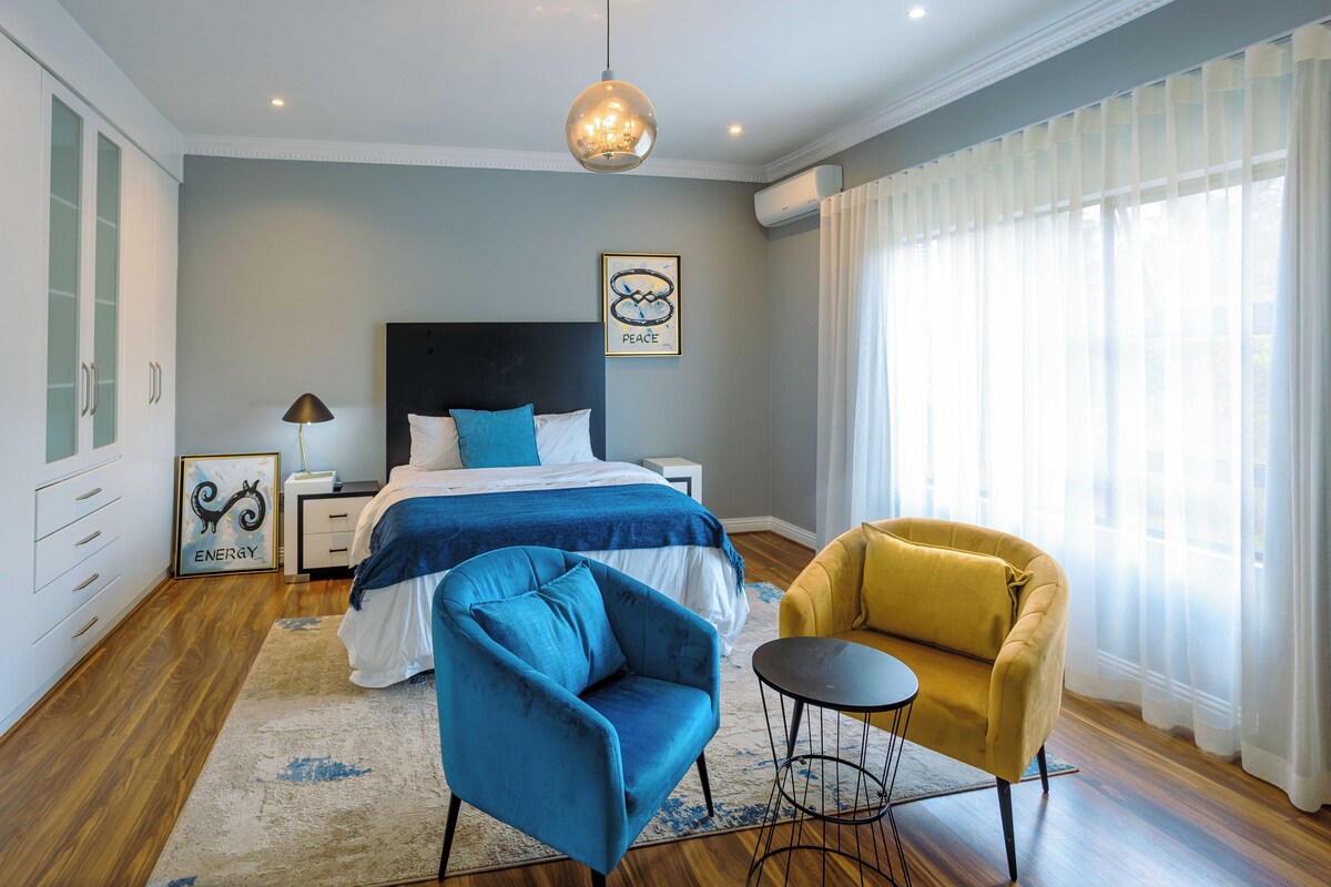 A bedroom with a bed, a blue armchair, a yellow armchair, a small round table, and a chandelier.