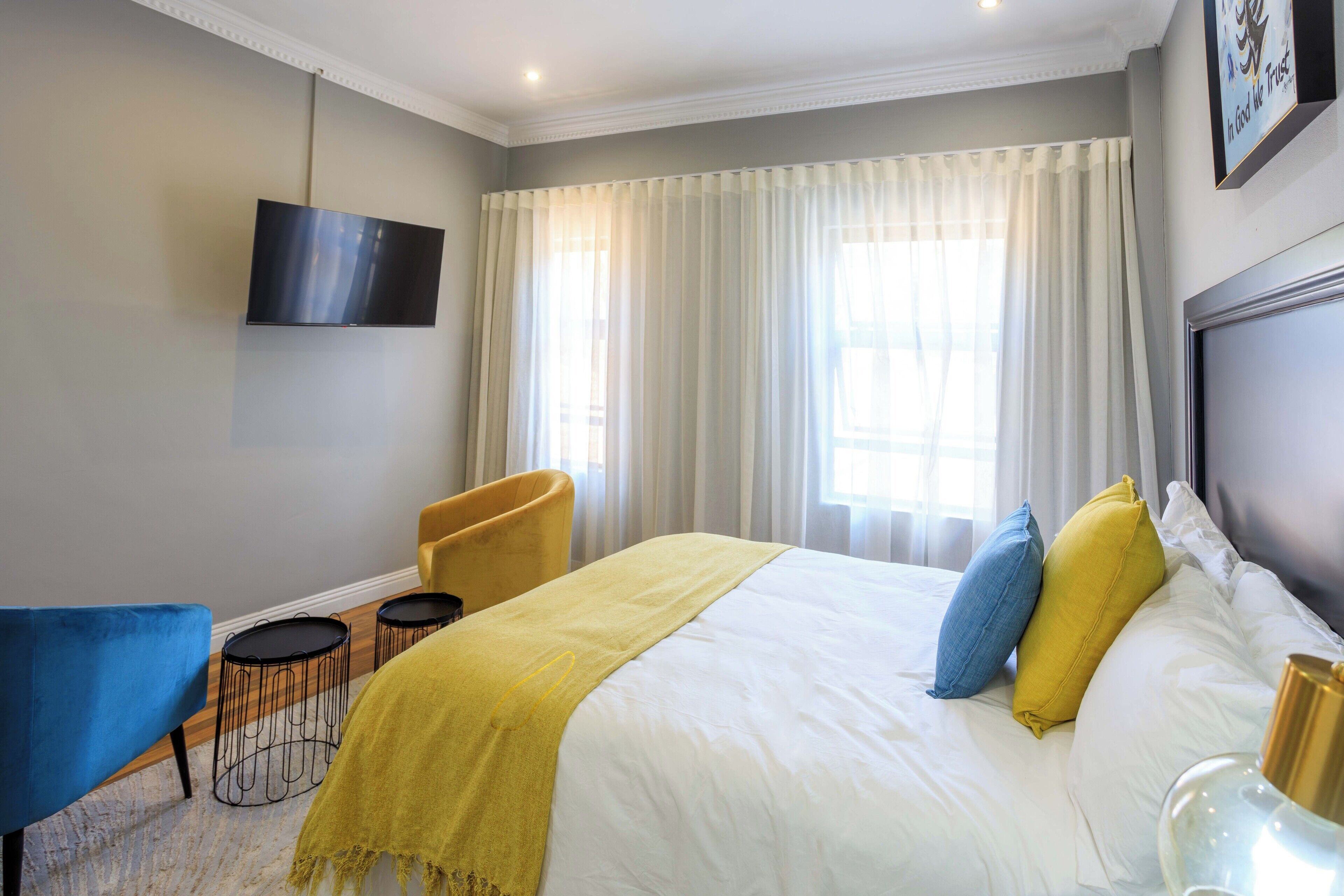 Habitación de hotel con cama, televisor montado en la pared, ventana con cortinas, un sillón amarillo y una mesita.