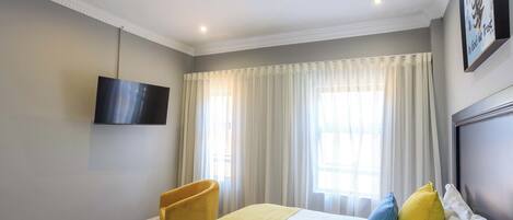 Habitación de hotel con cama, televisor montado en la pared, ventana con cortinas, un sillón amarillo y una mesita.