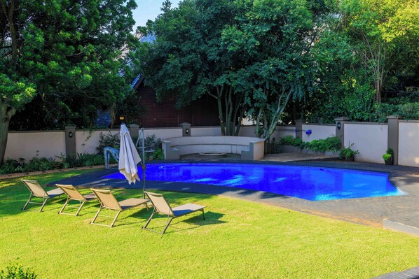 A swimming pool with lounge chairs and a covered seating area surrounded by trees and a brick wall. - Adinkra Boutique Hotel (Randburg)