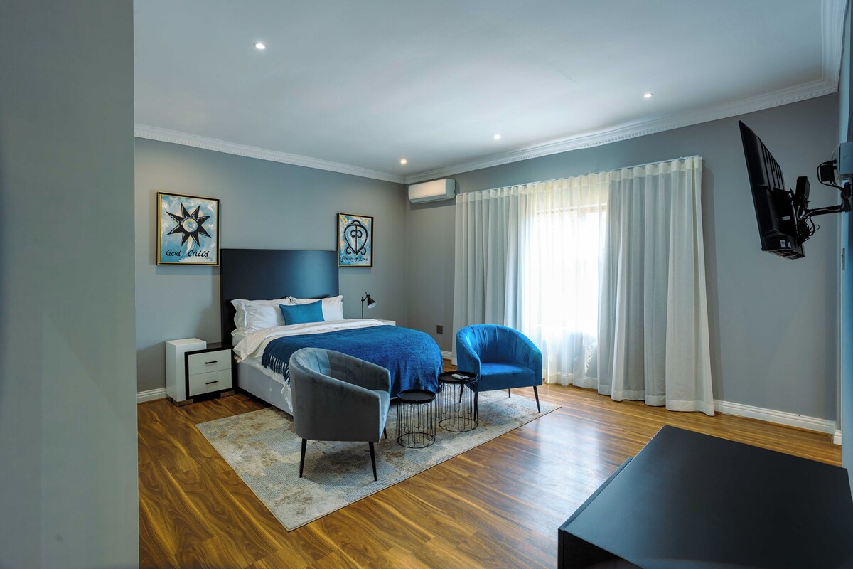 A modern bedroom with a bed, two chairs, a desk, and a flat-screen TV.