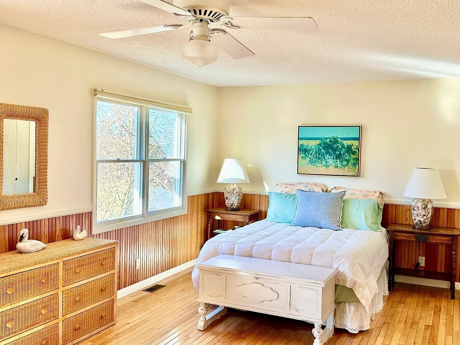 A bedroom with a bed, two bedside tables, a nightstand, a dresser, a ceiling fan, and a window with a view of trees.