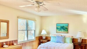 Une chambre à coucher comprenant un lit, deux tables de chevet, une commode, une coiffeuse, un ventilateur de plafond et une fenêtre donnant sur des arbres.