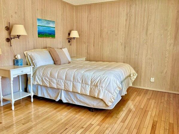 A bedroom with a wooden floor, a bed with a white duvet, two bedside lamps, a nightstand, and a painting on the wall.