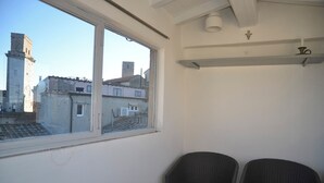 Ein Zimmer mit zwei Korbstühlen, einem Fenster mit Blick auf eine Stadtlandschaft mit Gebäuden und einem Turm, sowie einer weißen Decke mit Kreuz und einer Leuchte.