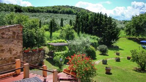 A stone building with a wooden staircase, a well-maintained garden with various plants and a red flowering bush, and a clear view of a forested hill in the background. - Farmhouse in Val D'orcia Charm (Castiglione d'Orcia)