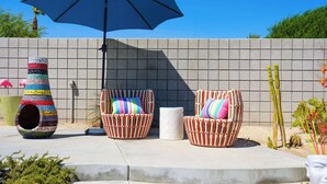 Een terras met twee rieten stoelen, een kleurrijke vogelhuisje en een blauwe parasol.