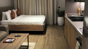 Ein modernes Hotelzimmer mit Bett, einer kleinen Couch, einem Couchtisch, einer Küchenzeile inklusive Spüle und Herd sowie einem großen Fenster mit Vorhängen.