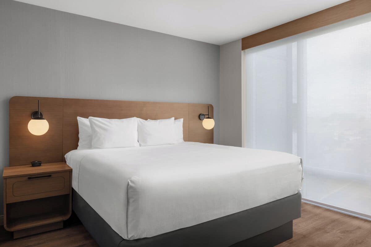 Ein modernes Hotelzimmer mit einem großen Bett, einem hölzernen Kopfteil, zwei Nachttischen mit Lampen und einem großen Fenster mit leichtem Vorhang.