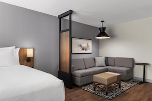 Ein modernes Hotelzimmer mit einem Bett, einem grauen Ecksofa, einem kleinen Tisch, einer Stehlampe und einem gerahmten Kunstwerk an der Wand.