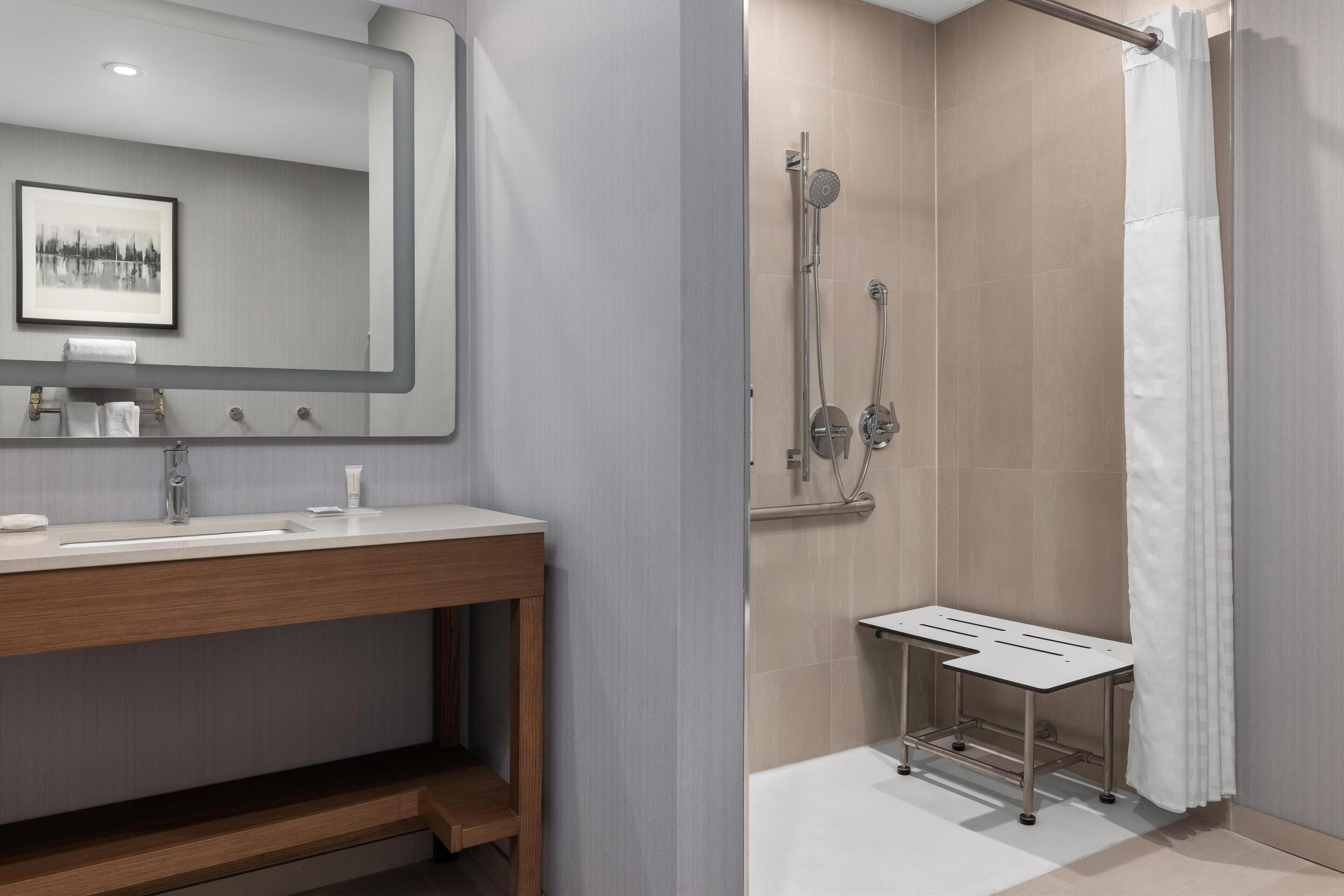 A modern bathroom with a large mirror, a wooden vanity, a shower with a handheld showerhead, and a white shower bench.