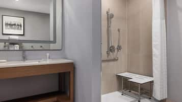 A modern bathroom with a large mirror, a wooden vanity, a shower with a handheld showerhead, and a white shower bench.