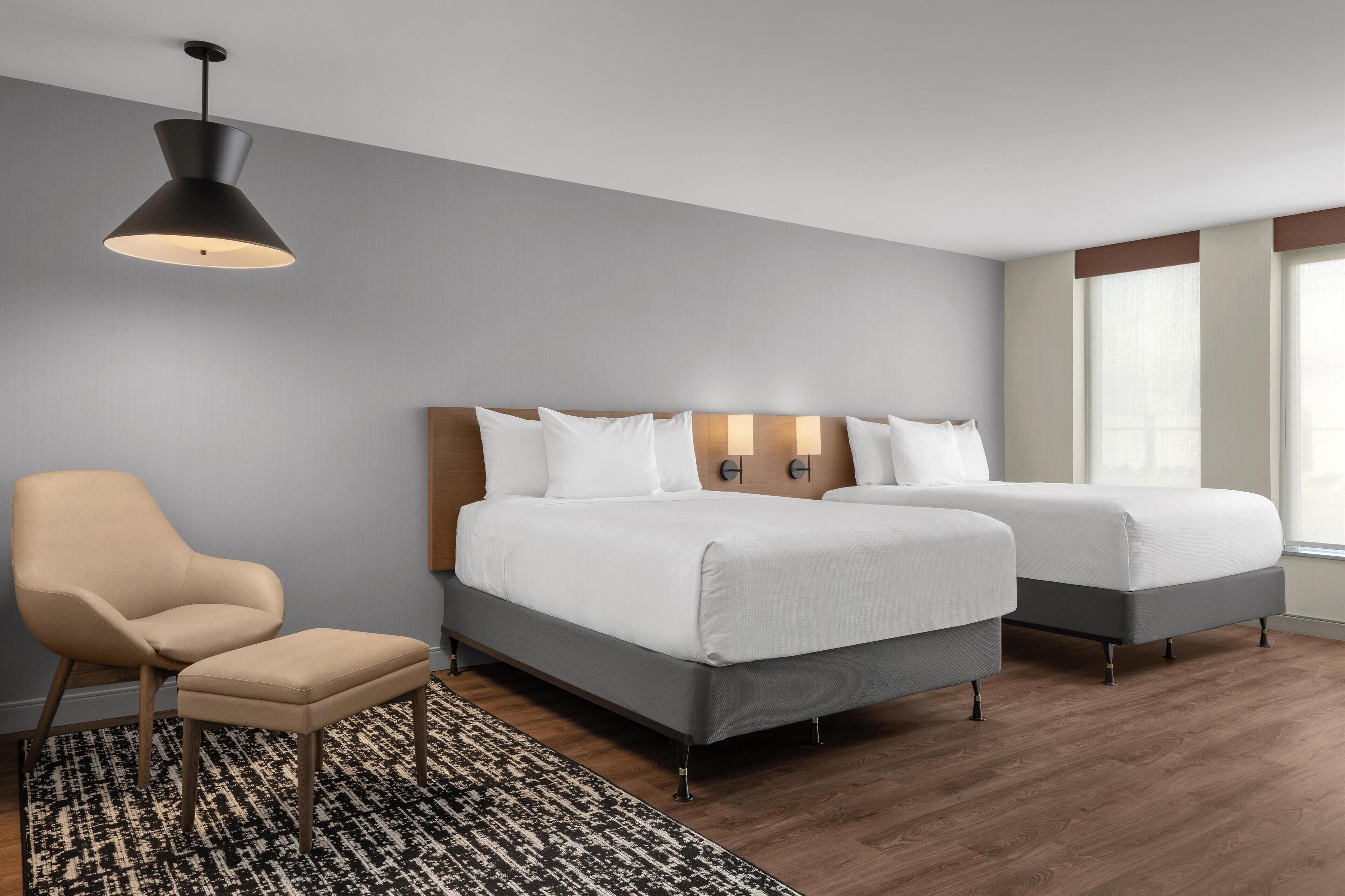 A hotel room with two beds, a chair, a small table, a patterned rug, and a hanging light fixture.