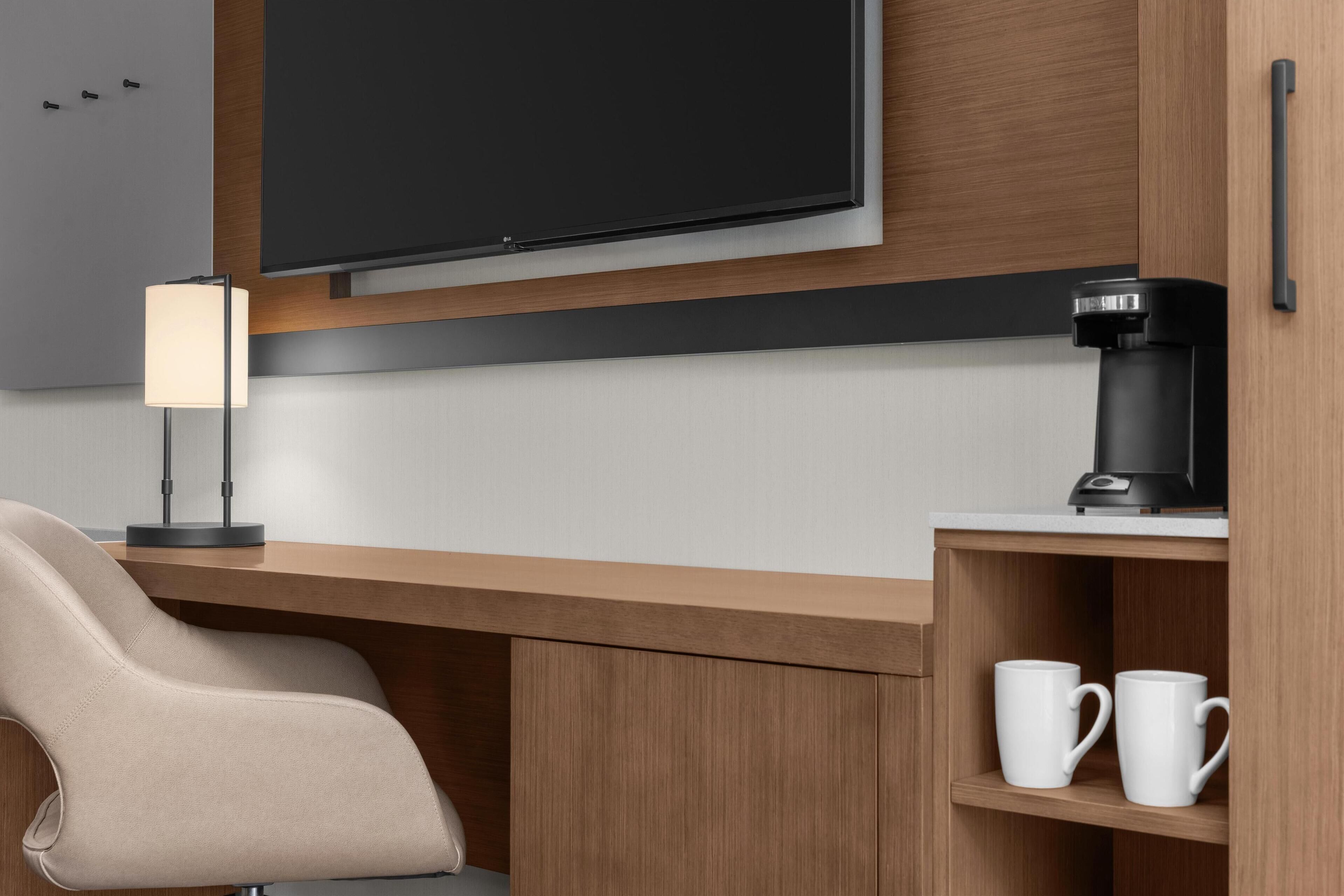 Una habitación de hotel moderna con un escritorio de madera, un televisor de pantalla plana, una lámpara, una cafetera y dos tazas blancas.