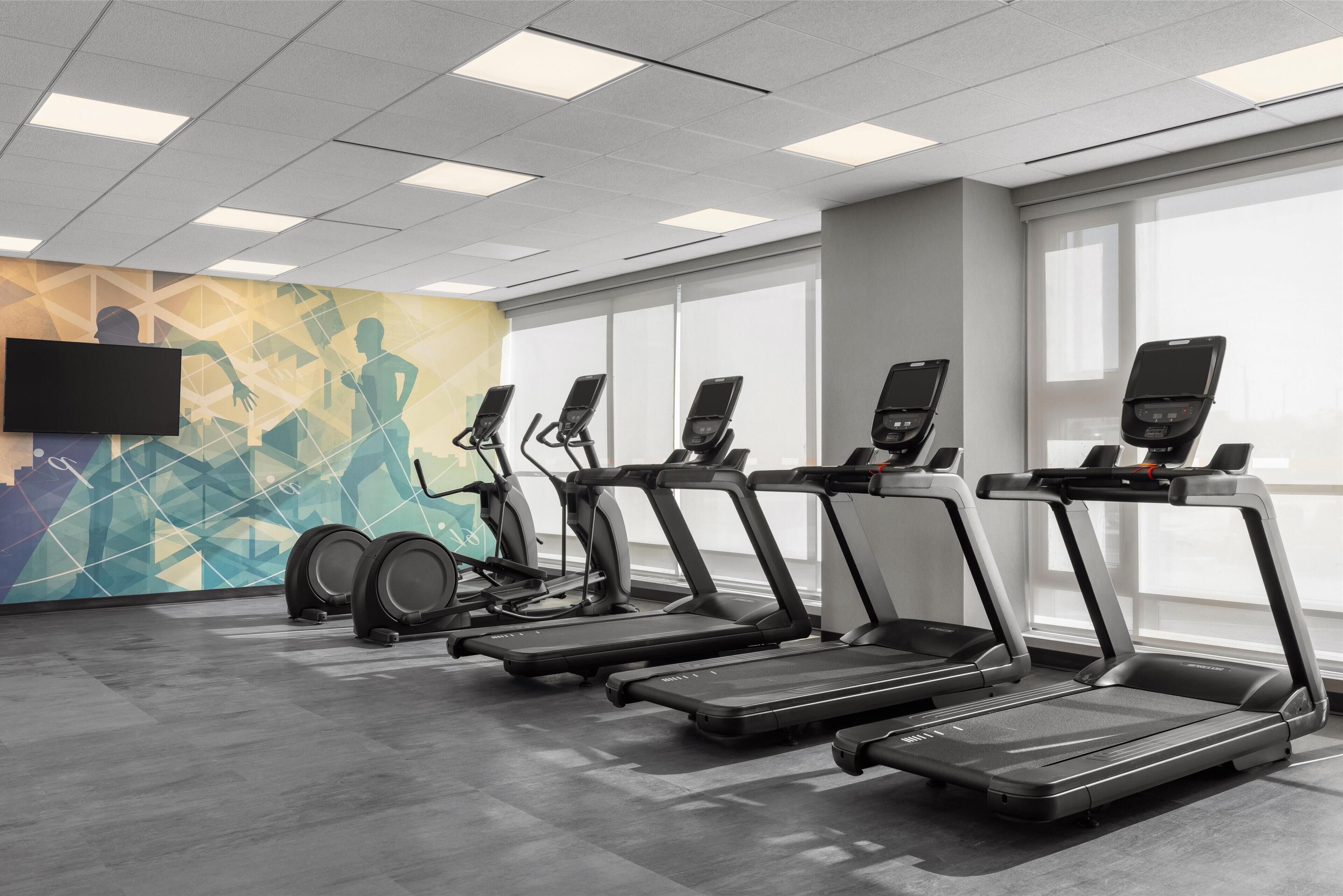 A gym with treadmills, stationary bikes, and a TV screen on the wall.