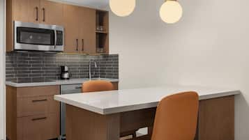 A modern kitchen with a built-in microwave, coffee maker, and a sink. There are wooden cabinets and a white countertop. Two orange chairs are placed at a small dining table.