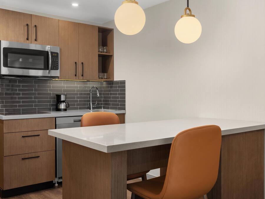 A modern kitchen with a built-in microwave, coffee maker, and a sink. There are wooden cabinets and a white countertop. Two orange chairs are placed at a small dining table.