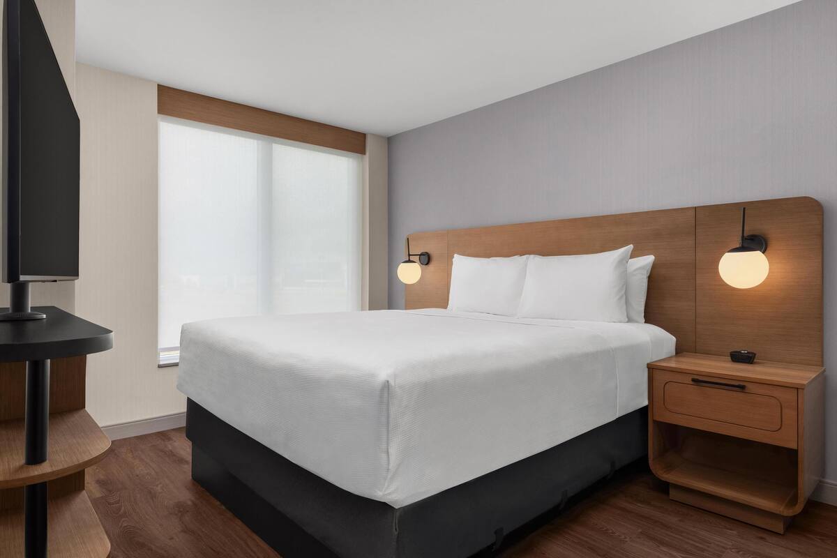 A modern hotel room with a large bed, wooden headboard, bedside tables, and a TV on a stand.