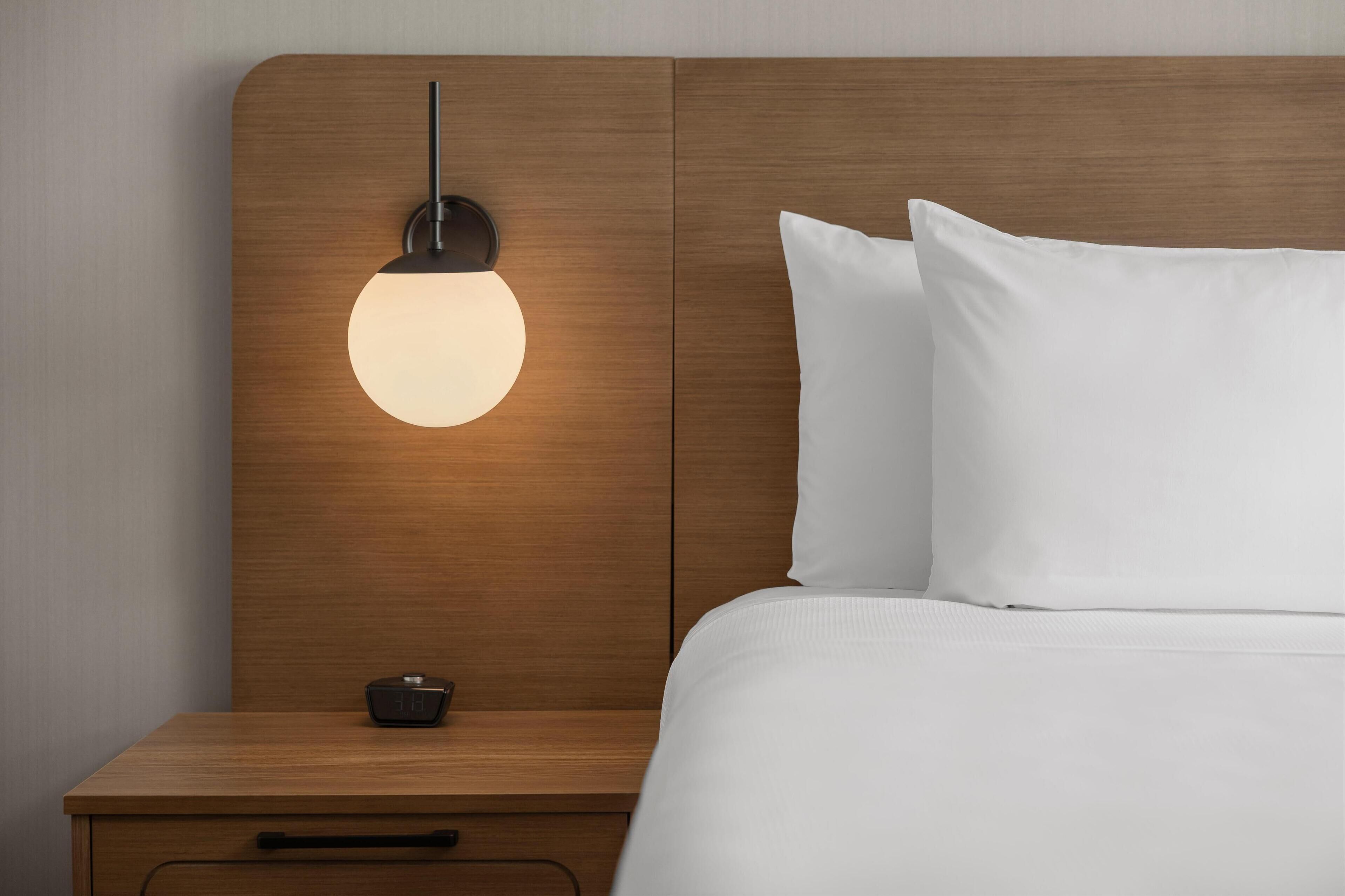 Una cama bien hecha con sábanas blancas, cabecera de madera y mesita de noche con lámpara.