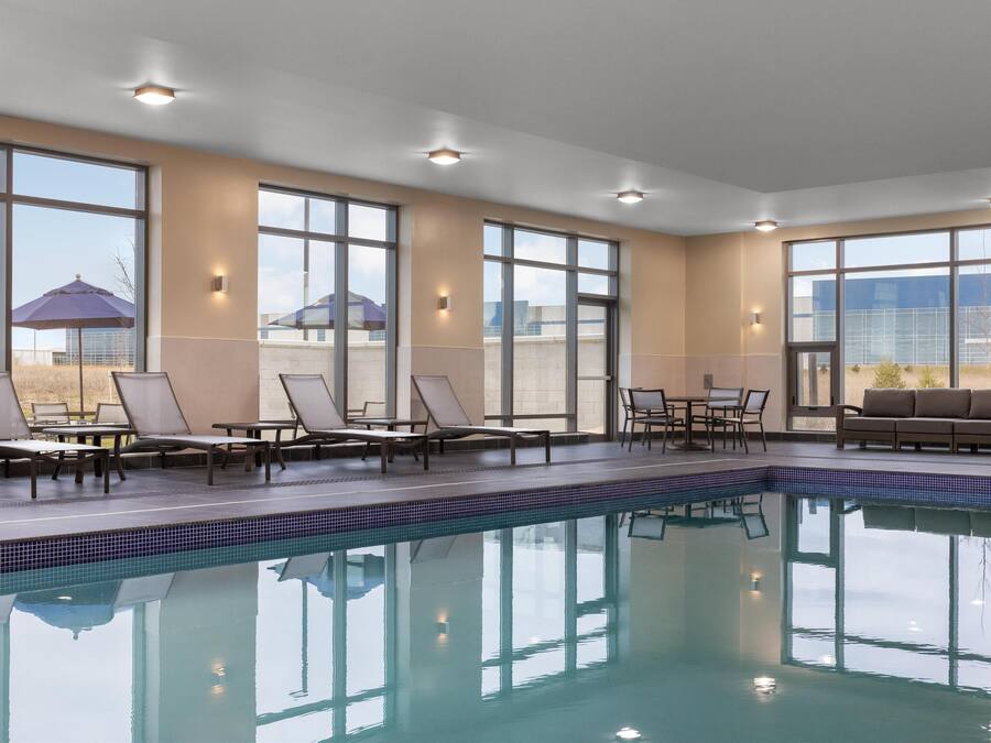 An indoor swimming pool with lounge chairs and tables, large windows, and a view of the outside.