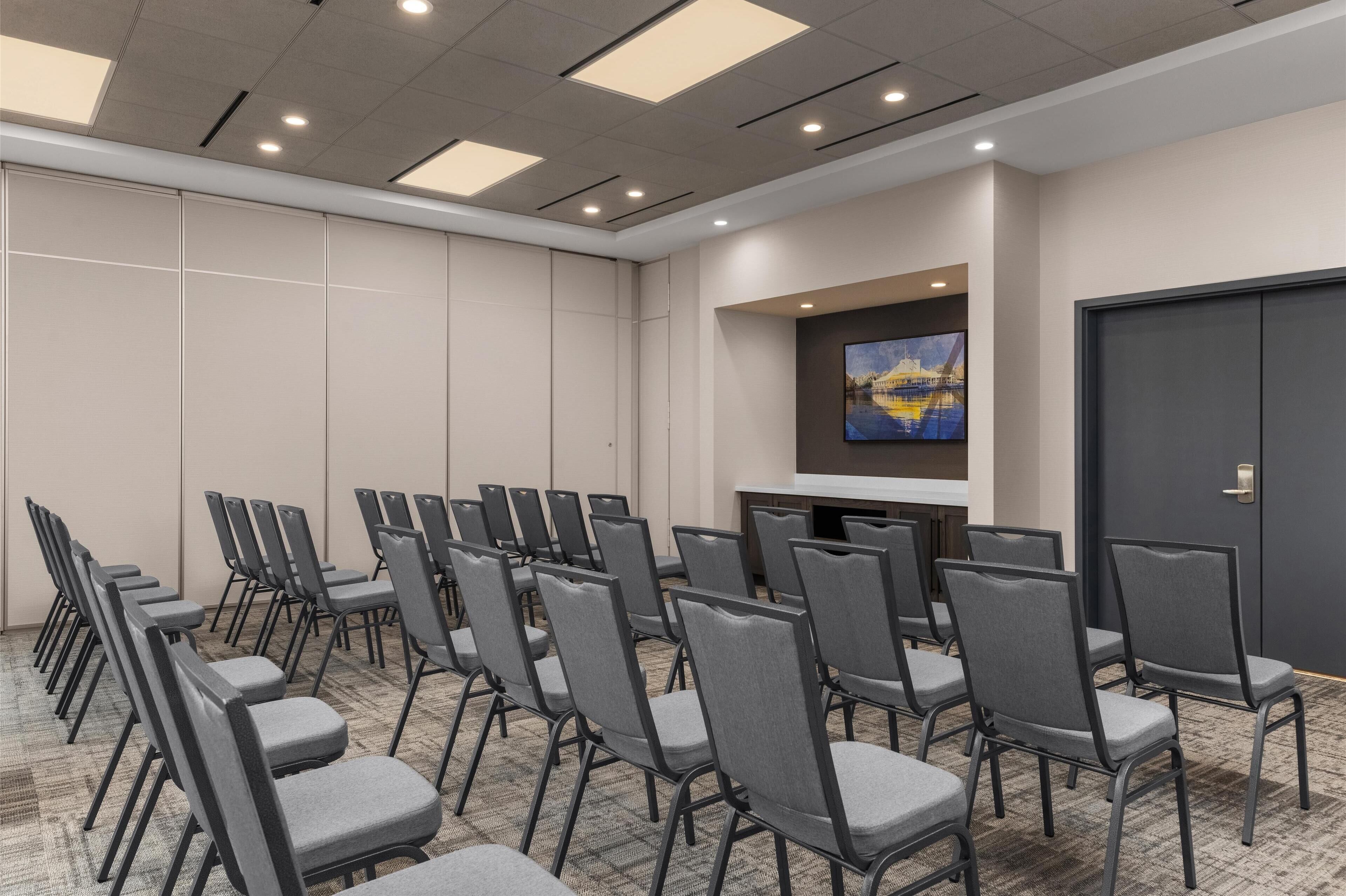 A conference room with rows of chairs, a flat-screen TV on the wall, and a door.