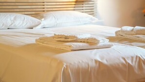 Una cama bien tendida con sábanas blancas y toallas dobladas, un cabecero de madera y una ventana con persianas.