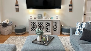 Een woonkamer met een grote flatscreen televisie aan de muur, grijze banken, een bijzettafeltje met dienblad en decoratieve objecten.
