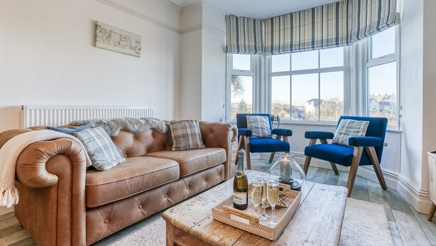 Ein Wohnzimmer mit einem braunen Ledersofa, zwei blauen Sesseln, einem Holztisch mit einer Flasche und Gläsern sowie Blick auf Häuser durch große Fenster.