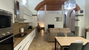Eine moderne Küche mit Einbauofen, Mikrowelle und Dunstabzugshaube aus Edelstahl. Es gibt einen Essbereich mit Holztisch und Stühlen sowie ein Wohnzimmer mit Sofa und Fernseher.
