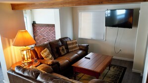 A living room with a brown leather sofa, a wooden coffee table, a lamp, and a flat-screen TV mounted on the wall. -  Escape to a cozy cottage on Ausable Lake, in Lupton, Michigan! (Lupton)