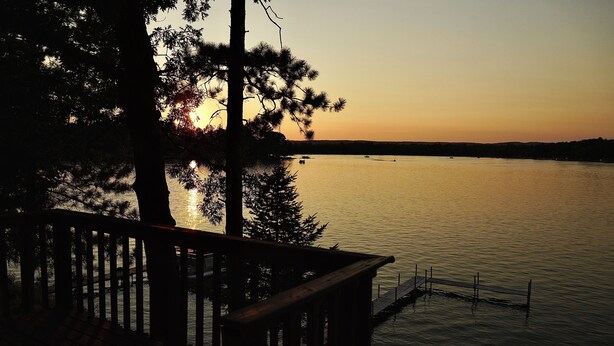 A lakeside view with a wooden deck and dock extending into calm water, surrounded by trees. -  Escape to a cozy cottage on Ausable Lake, in Lupton, Michigan! (Lupton)