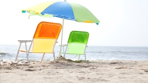 Twee strandstoelen onder een kleurrijke parasol op een zandstrand, met de oceaan op de achtergrond.