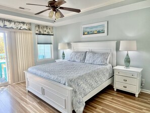 A bedroom with a bed, two bedside tables with lamps, a ceiling fan, a window with blinds, and a painting on the wall.