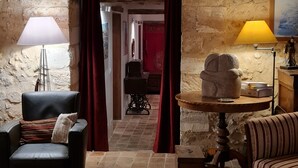 A rustic room with stone walls, a leather chair, a wooden table with a lamp, and a patterned armchair. - DOMAINE DE LABROUSSE (Agonac)