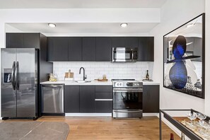 A modern kitchen with dark cabinets, stainless steel appliances, a white tiled backsplash, and a cityscape artwork on the wall.