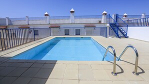 En swimmingpool med ett metallräcke, en kakelbeklädd terrass och en trappa som leder upp till en byggnad med en vit fasad och flera fönster.