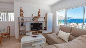 A modern living room with a large window, a sofa, a coffee table, and a TV stand with decorative items. - Blue Villa Rest & Pool (Velilla-Taramay)
