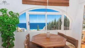 A dining area with a round wooden table and cushioned chairs, a potted plant, and a view of the sea through a large arched window. - Blue Villa Rest & Pool (Velilla-Taramay)
