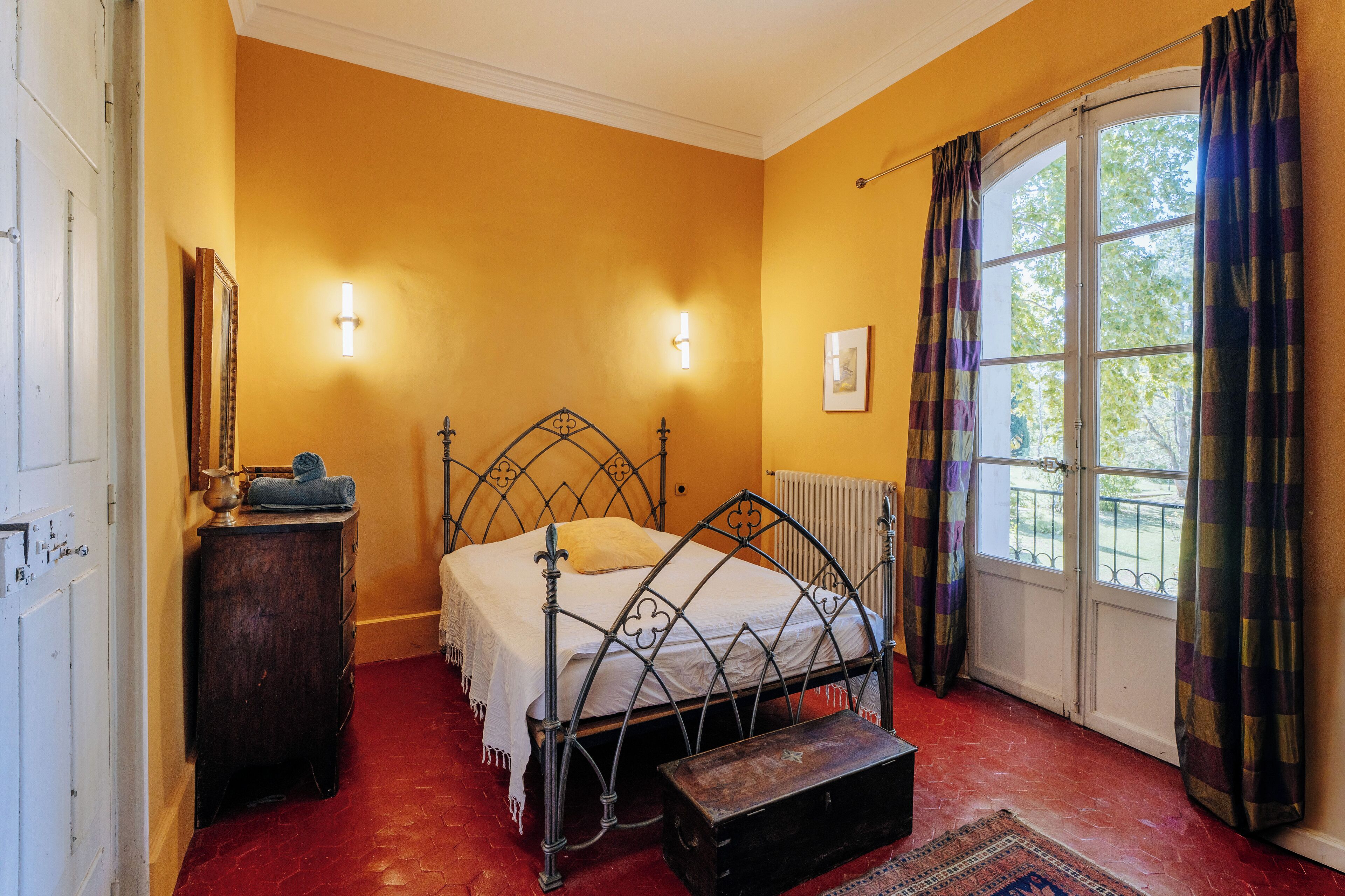 Quarto com estrutura de cama de metal, cômoda de madeira, um banco e janela com cortinas.