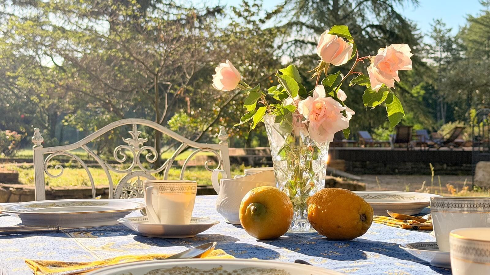 A table set outdoors with a vase of flowers, a teapot, cups, and oranges.
