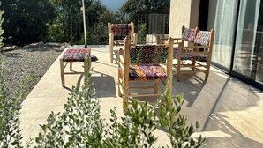 An outdoor seating area with wooden chairs and a table, covered by a large umbrella, with a view of trees and mountains in the background.