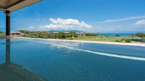 Vista despejada de una piscina infinita con cubierta de madera, que se extiende hacia un paisaje verde exuberante con un campo de golf y el océano al fondo.