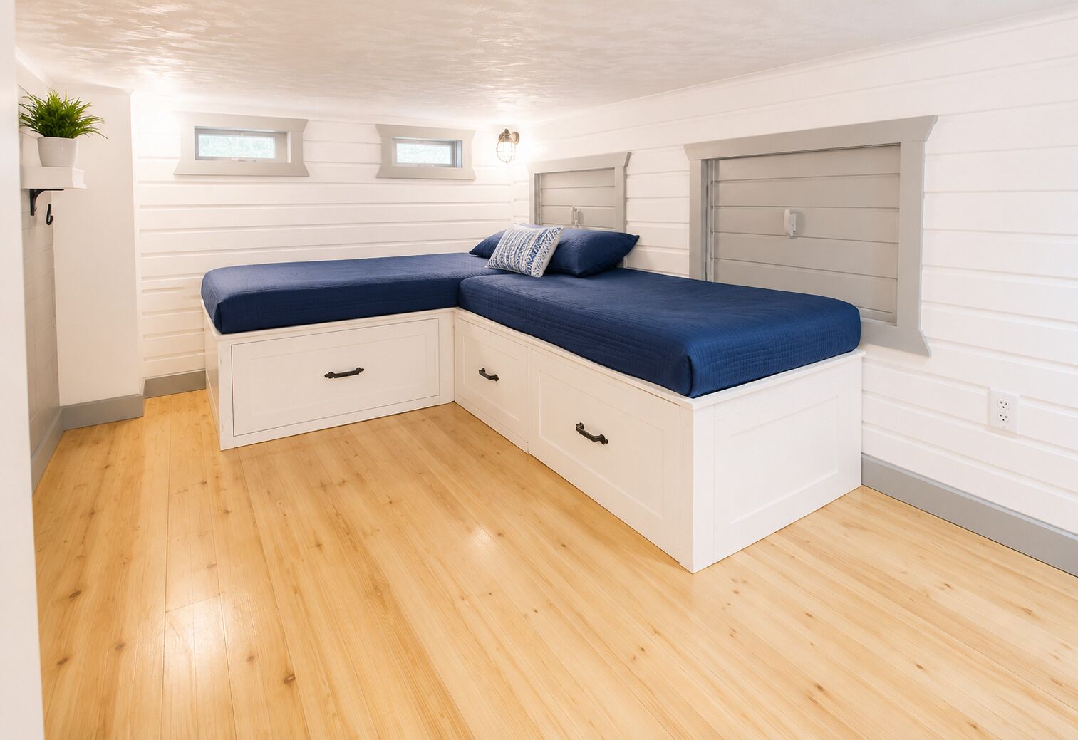 Une chambre d’angle avec un lit intégré, deux tiroirs, une fenêtre et une plante.