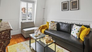 A living room with a black leather sofa, a glass coffee table, and a grey armchair. There is a window with a view of buildings outside. - Arc de Triomphe - Lauriston 1bdr by B'Your Home (Paris)
