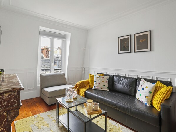 A living room with a black leather sofa, a glass coffee table, and a grey armchair. There is a window with a view of buildings outside.